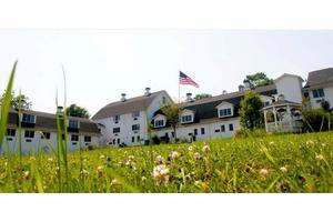 A large farmhouse with an American flag, surrounded by a grassy field on a sunny day.