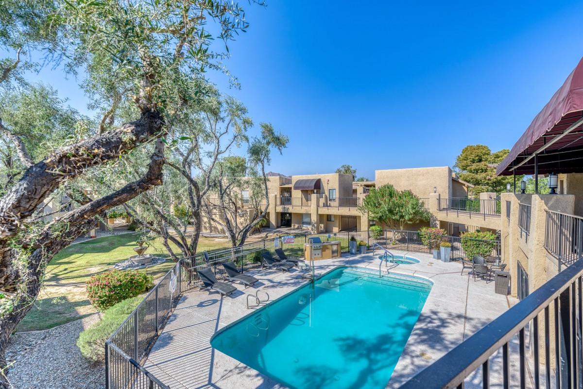 A sunny view of an apartment complex featuring a well-maintained outdoor swimming pool surrounded by trees.