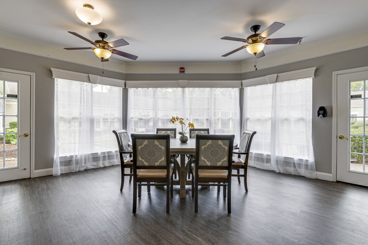 A spacious dining room with a round table, surrounded by chairs and large windows covered with sheer curtains.