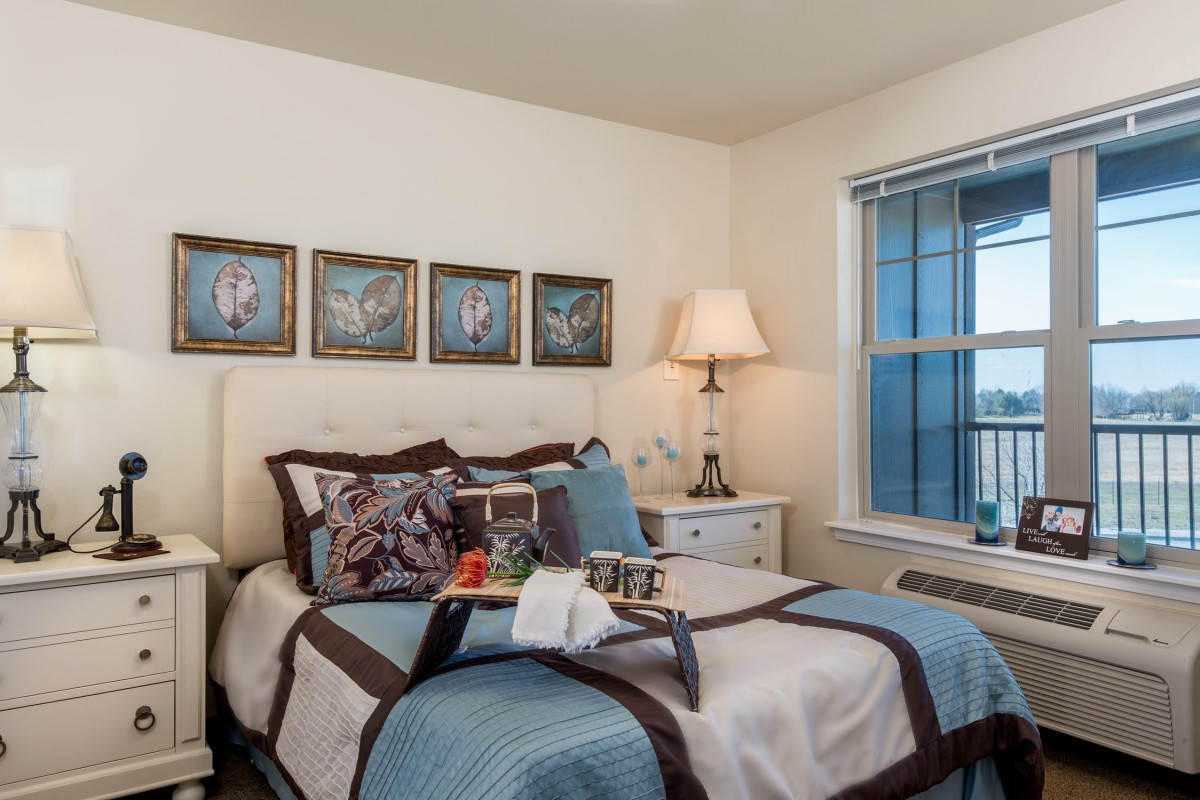 A cozy bedroom with a neatly made bed, decorative pillows, a bedside lamp, and framed artwork above the headboard.