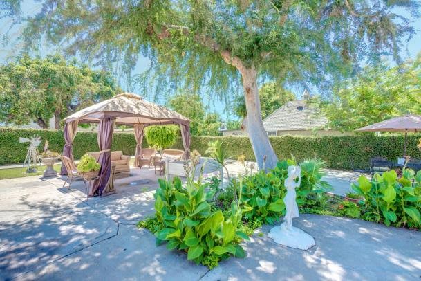 A serene garden with a gazebo, surrounded by lush greenery and a sculpture under a sunny sky.