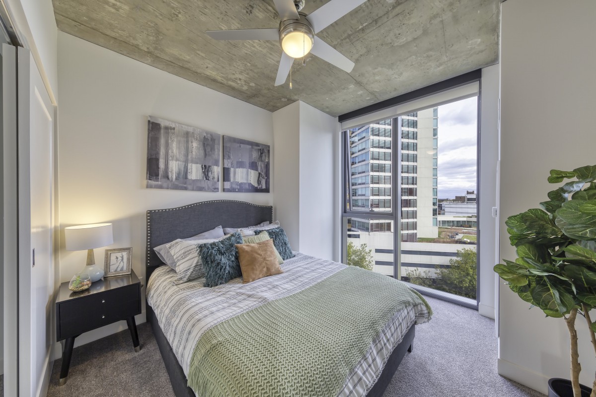 A modern bedroom with large windows offering a city view, featuring contemporary decor and ample natural light.