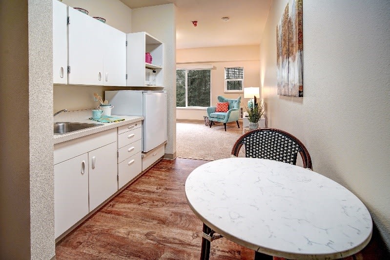 A small kitchen with white cabinets opens up to a cozy living area featuring wooden floors and modern decor.