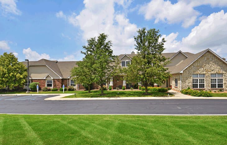 A well-maintained residential house with a green lawn and surrounding trees under a partly cloudy sky.