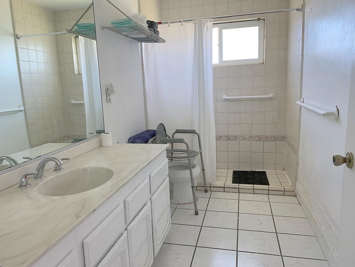 A clean, white-tiled bathroom with a sink, mirror, and accessible shower area.