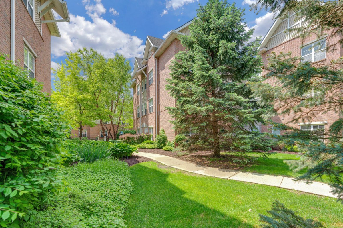 A scenic view of a well-maintained pathway lined with lush greenery and trees, next to a residential building under a partly cloudy sky.