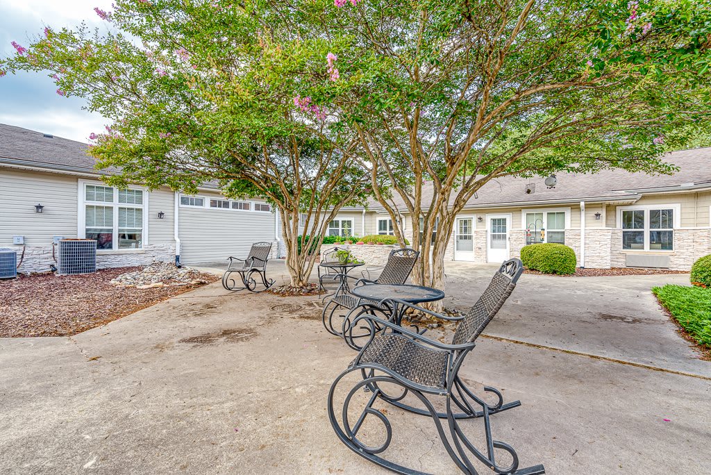 A peaceful outdoor patio featuring rocking chairs and tables under a tree, set in a courtyard with surrounding buildings.