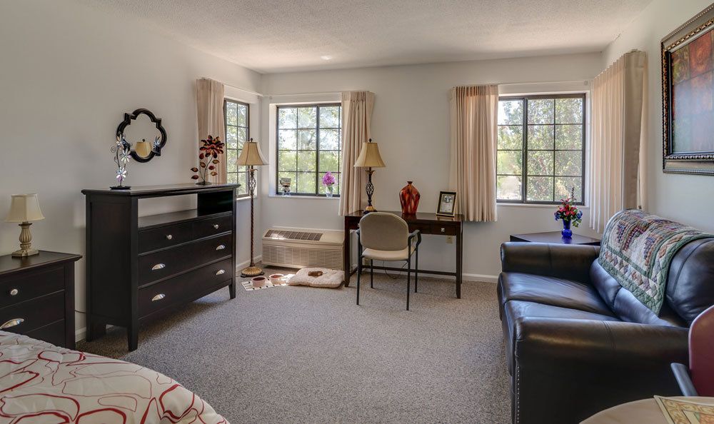 A cozy bedroom featuring a desk, chair, dresser, and windows with curtains.