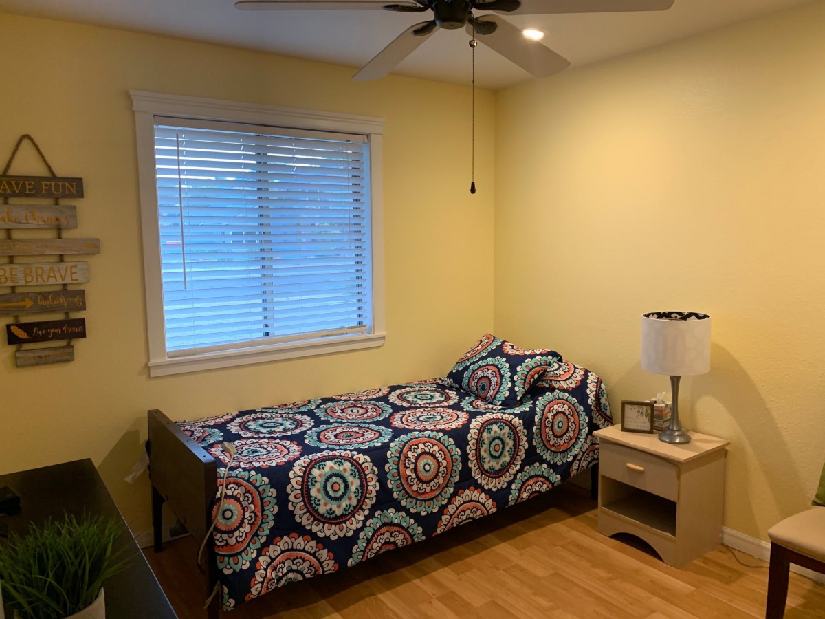 A cozy bedroom with a single bed featuring colorful bedding, a nightstand with a lamp, and a window illuminating the room.