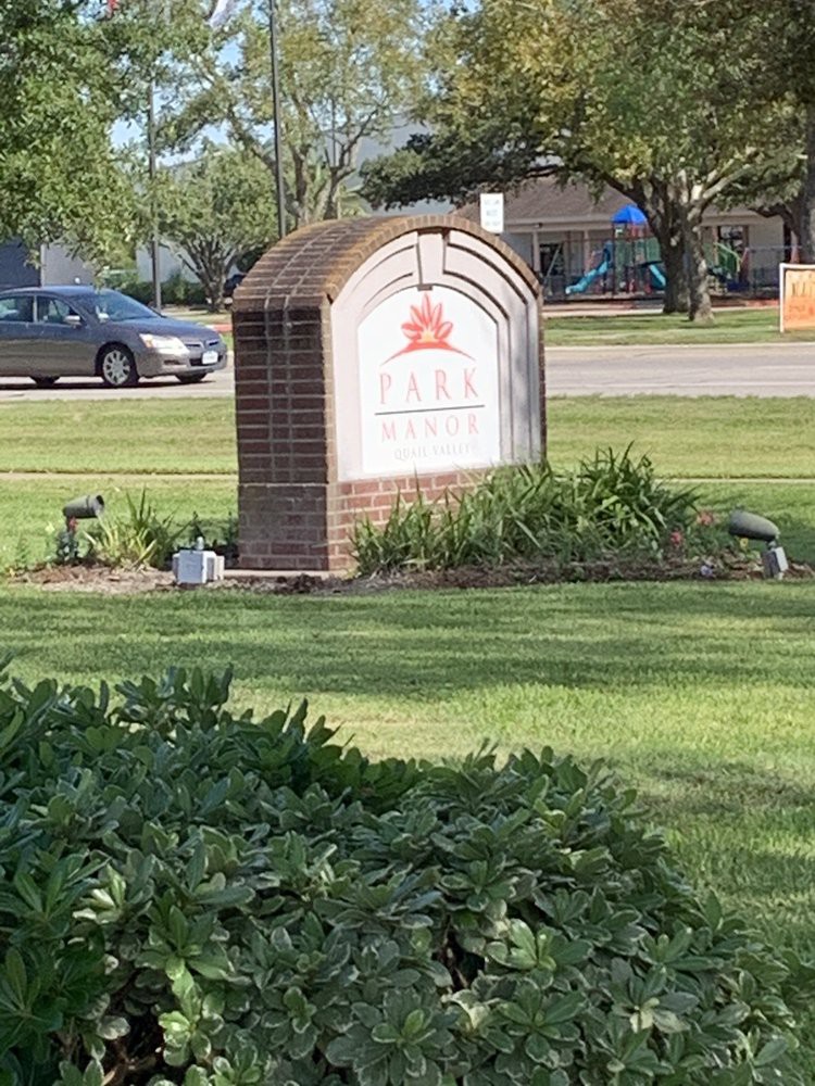 A park sign surrounded by greenery and a nearby road with a car.