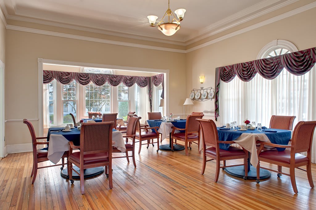 Elegant dining room with neatly arranged tables and chairs, featuring large windows with draped curtains.