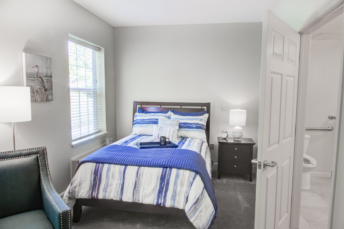 A cozy bedroom with a neatly made bed, featuring blue and white bedding, a nightstand with a lamp, and natural light coming through the window.