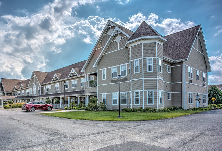A modern residential building with a neatly maintained lawn and parking area under a partly cloudy sky.