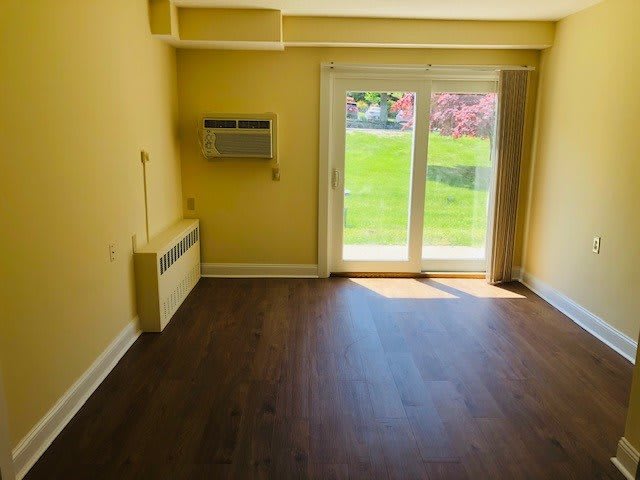 An empty room with wooden flooring and yellow walls, featuring an air conditioner and a sliding glass door leading to a green lawn.