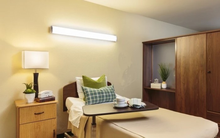 A clean and well-organized hospital room featuring a bed with green pillows, a wooden cabinet, and bright lighting.
