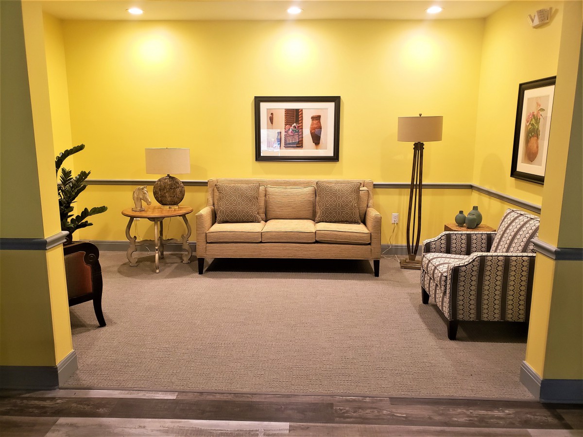 A cozy living room with modern furniture, featuring a sofa and armchair against a bright yellow wall adorned with artwork.