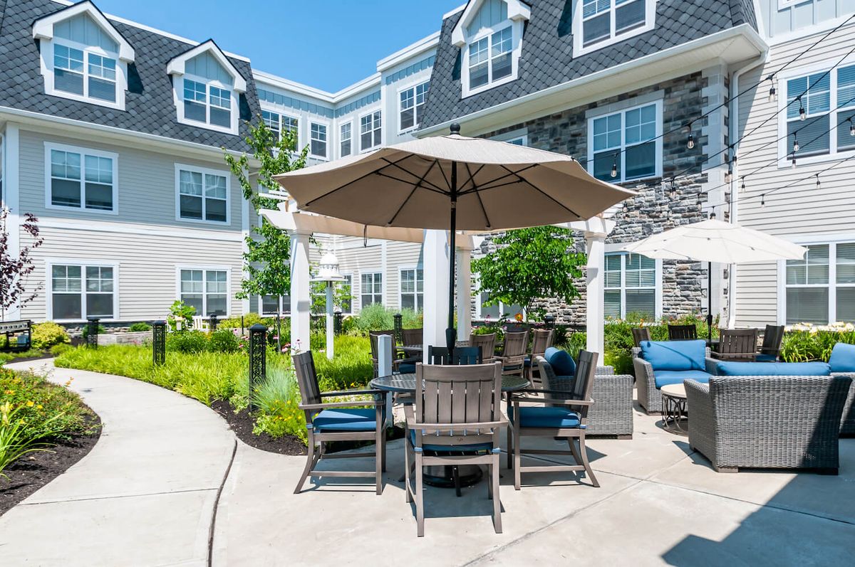 An outdoor patio area with a table and chairs under an umbrella, set in front of a residential building on a sunny day.