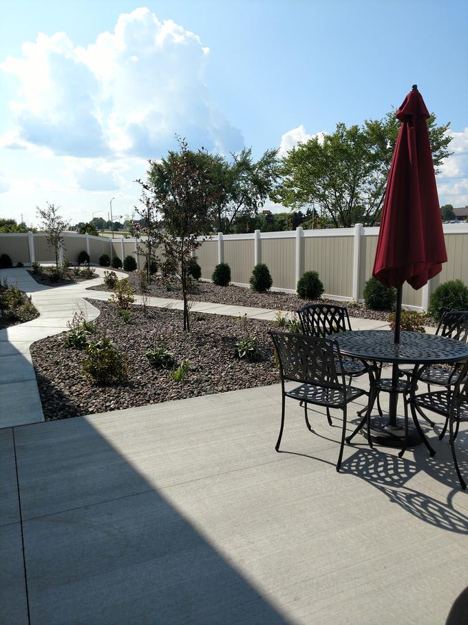 An outdoor patio with a table, chairs, and an umbrella, surrounded by landscaped areas and a pathway.