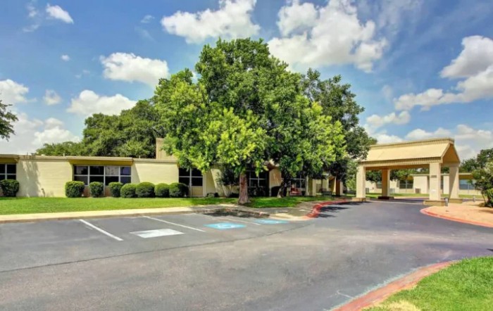 A sunny day view of a building surrounded by trees with an adjacent parking lot.