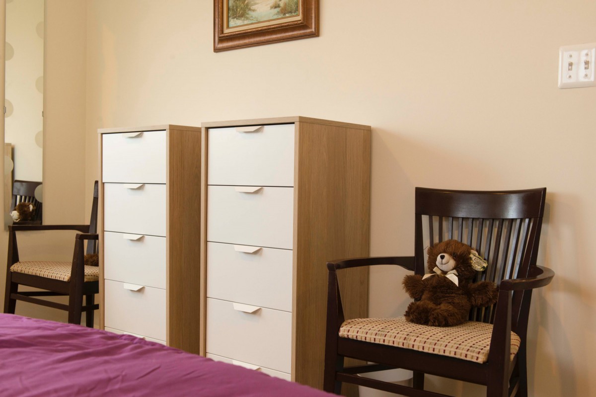 A cozy bedroom featuring a pair of dressers, a chair with a teddy bear, and a painting on the wall.