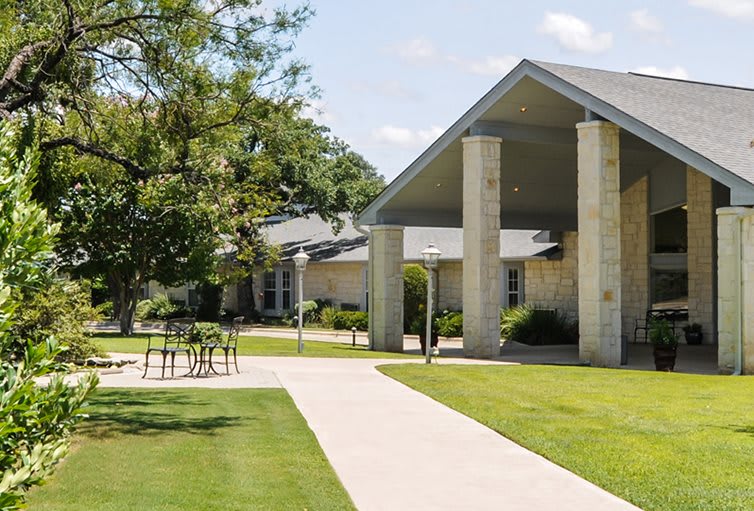 A modern building with a sloped roof and stone columns, surrounded by grassy areas and trees.