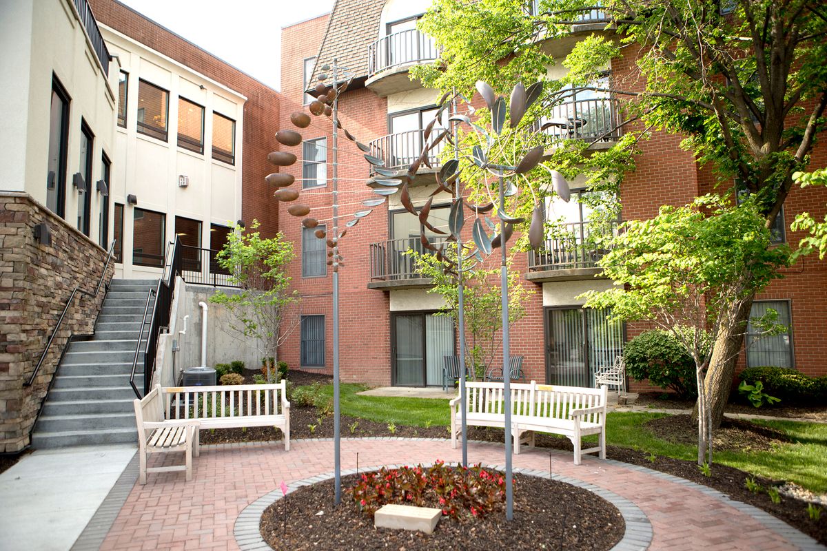 A tranquil courtyard featuring a central sculpture, surrounded by benches and greenery, adjacent to a brick building.