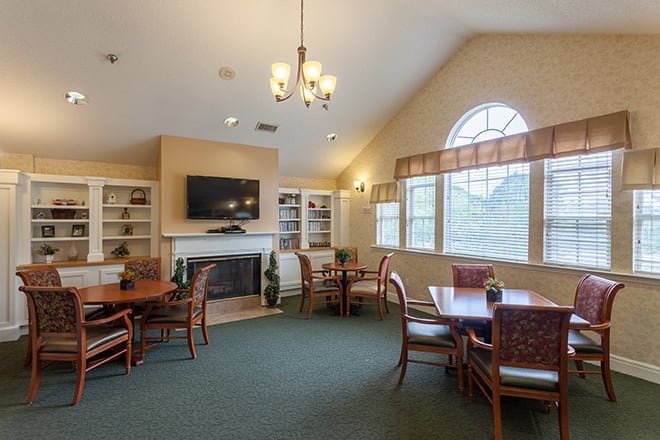 A cozy living room with multiple tables and chairs, a television above a fireplace, bookshelves on either side, and large windows providing natural light.