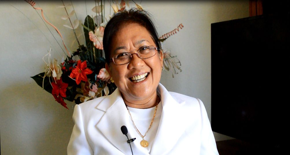A person wearing glasses and a white outfit, smiling with a microphone attached, standing in front of flowers.