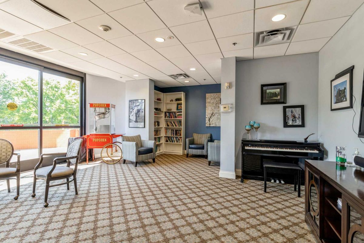 A cozy lounge area featuring comfortable chairs, a piano, a bookshelf, and large windows.