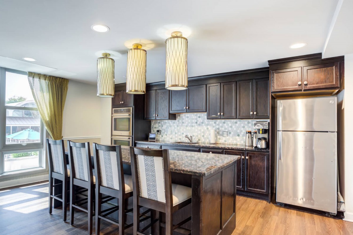 A modern kitchen featuring dark cabinets, a granite countertop, stainless steel appliances, and pendant lighting above the bar area with stools.