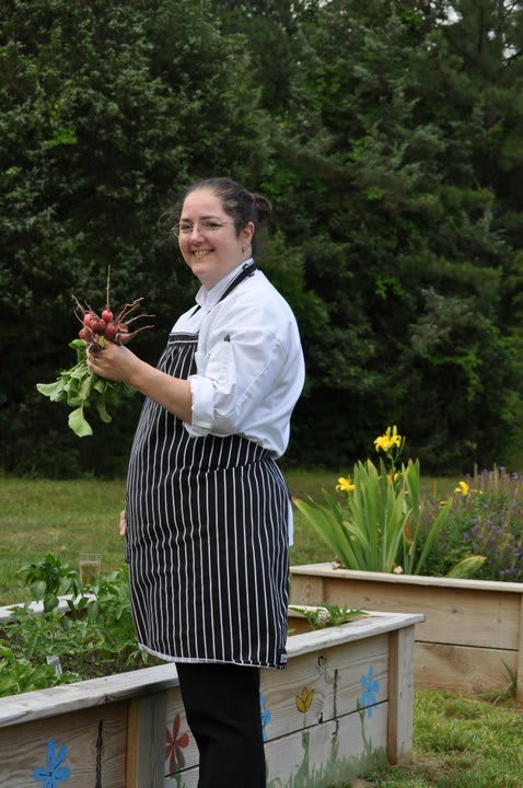 A chef in an apron holding freshly picked vegetables in a garden.
