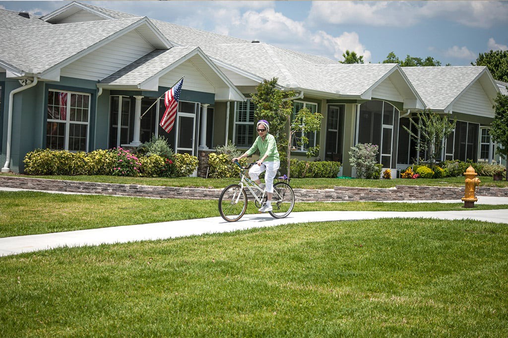 A person rides a bicycle along a sidewalk in a suburban neighborhood on a sunny day, with houses and an American flag in the background.