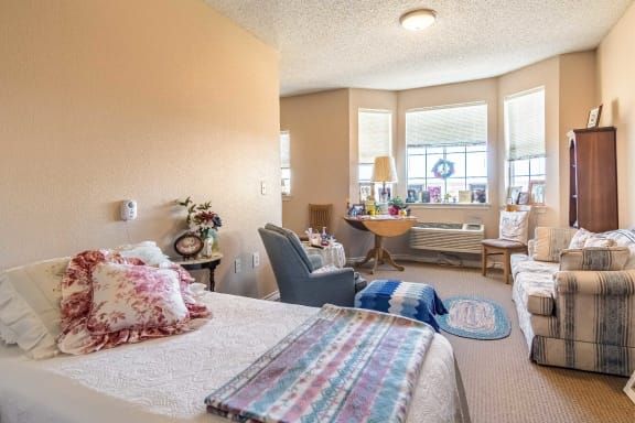 A cozy bedroom with ample sunlight streaming through large windows, featuring comfortable seating and homey decor.