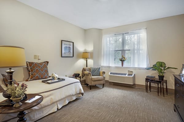 A cozy hotel bedroom with a neatly made bed, armchair, and sunlight streaming through the window.