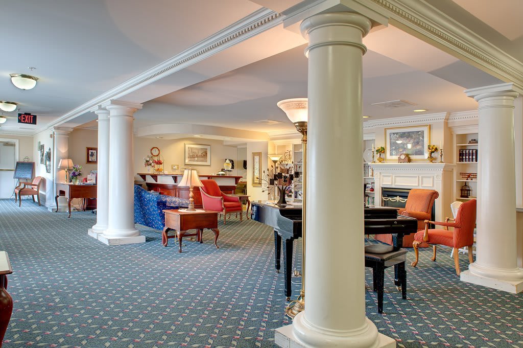 A spacious lounge area with elegant columns, a piano, and colorful chairs arranged on a patterned carpet.