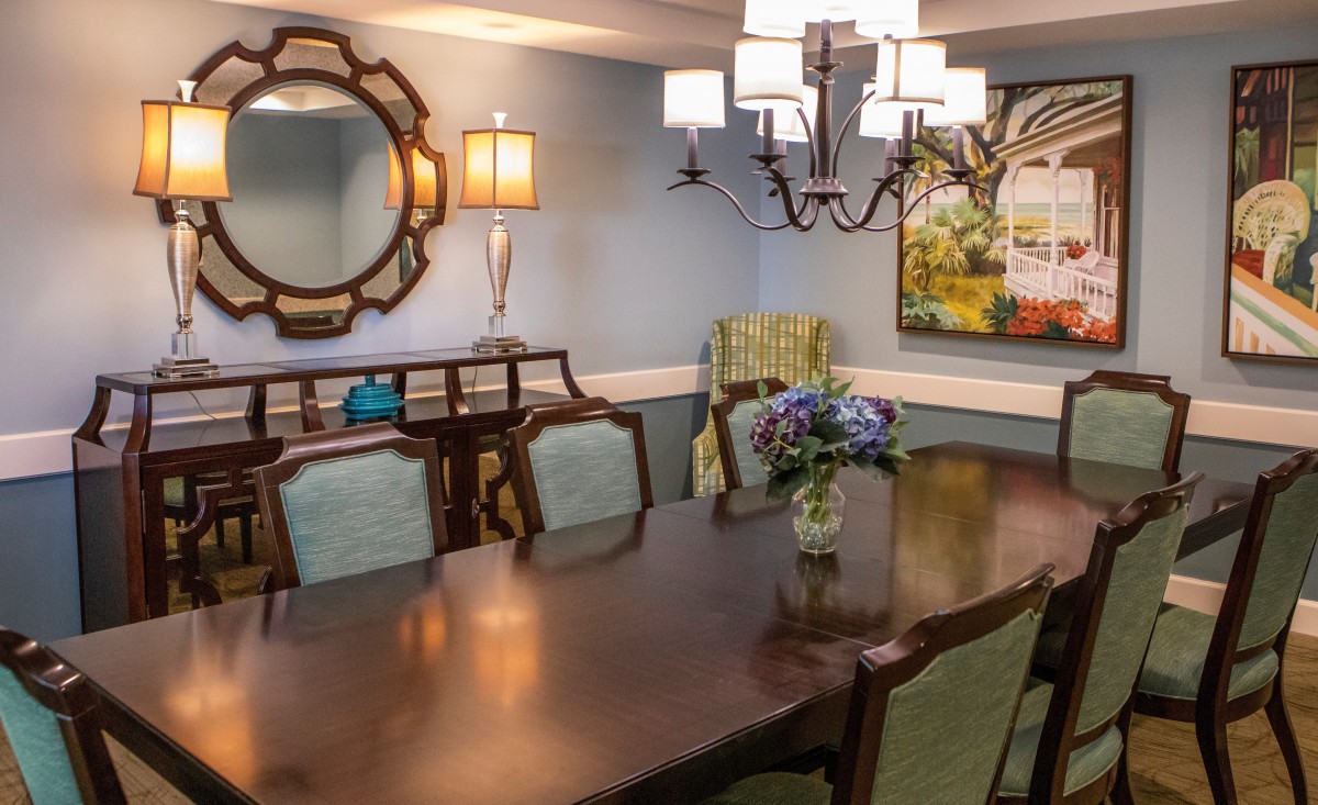A well-decorated dining room with a large wooden table, elegant chairs, a decorative mirror, chandelier lighting, and colorful artwork on the wall.