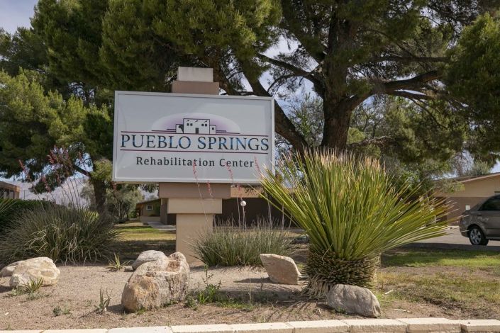 Sign for Pueblo Springs Rehabilitation Center surrounded by trees and plants.