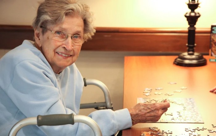 An elderly woman sits in a wheelchair, smiling as she works on a puzzle.