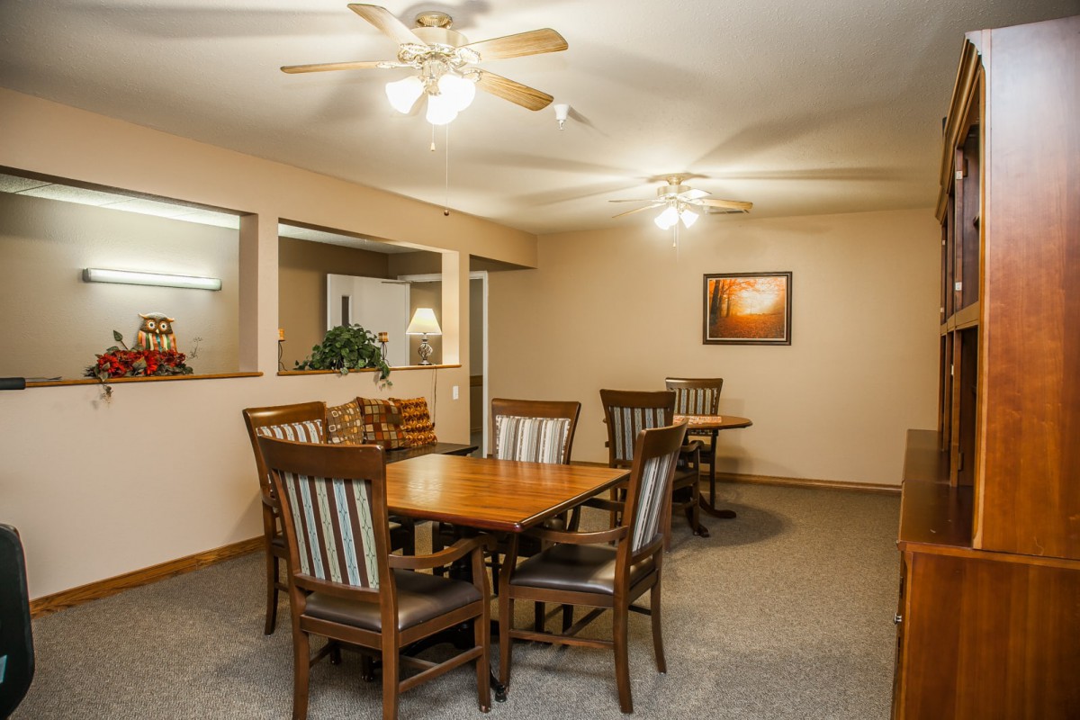 A dining room setting with wooden tables and chairs, ceiling fans, and a carpeted floor.