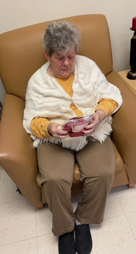 An elderly woman sits comfortably in a chair, engrossed in reading something while wrapped in a cozy blanket.