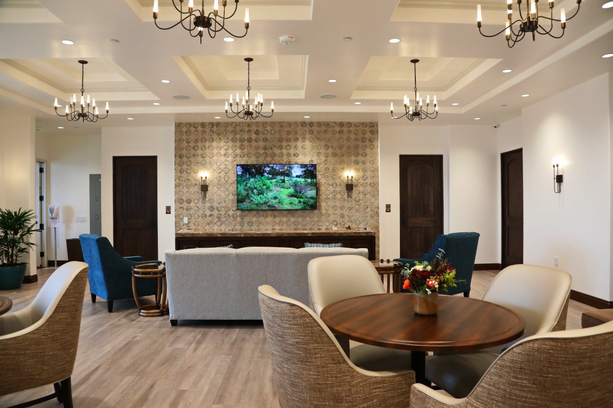 A modern living room featuring comfortable seating, a wall-mounted television, elegant chandeliers, and a cozy round table.