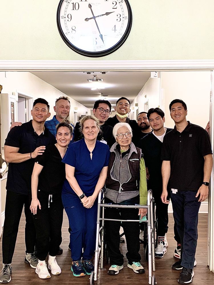 A group of healthcare workers stand with an elderly woman using a walker in a hospital hallway.