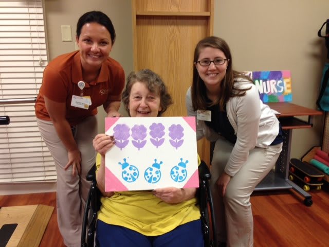 A smiling woman in a wheelchair proudly displays her artwork, accompanied by two women in a healthcare setting.