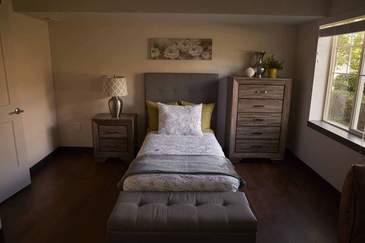 A cozy bedroom with a single bed, nightstand, lamp, and dresser in soft lighting.
