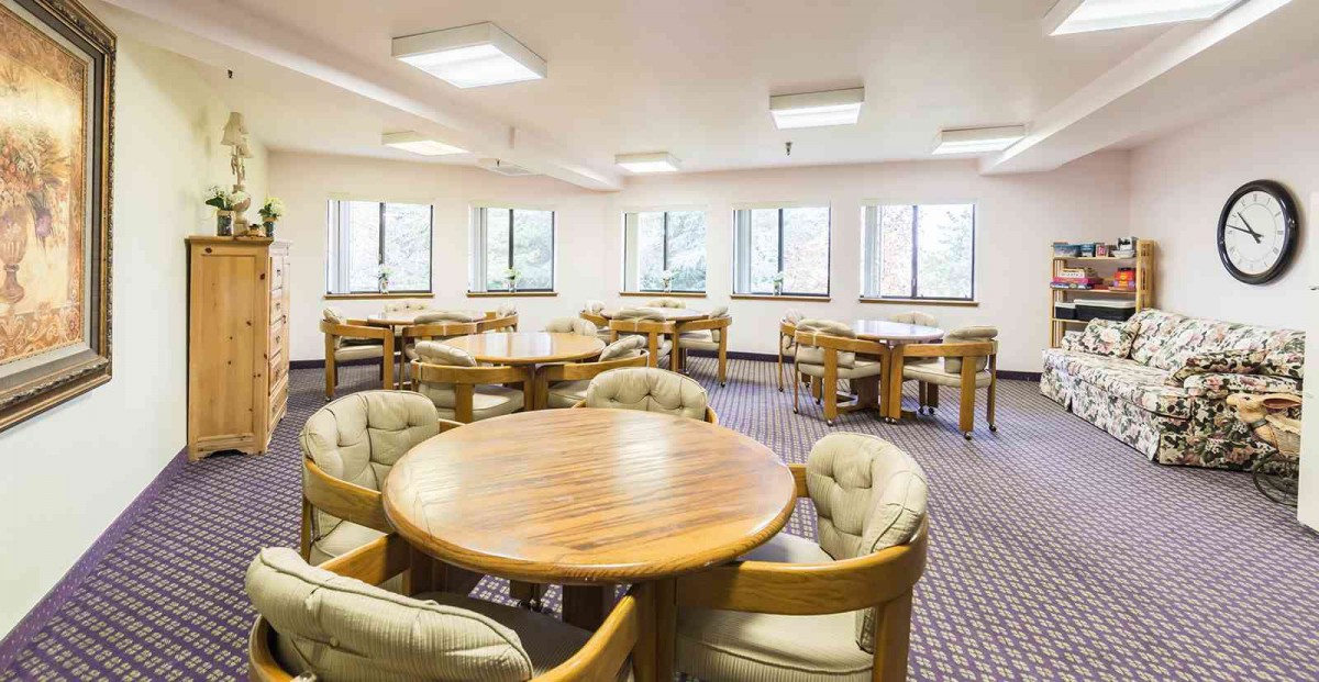 A spacious dining area with multiple tables and chairs, featuring large windows and carpeted flooring.