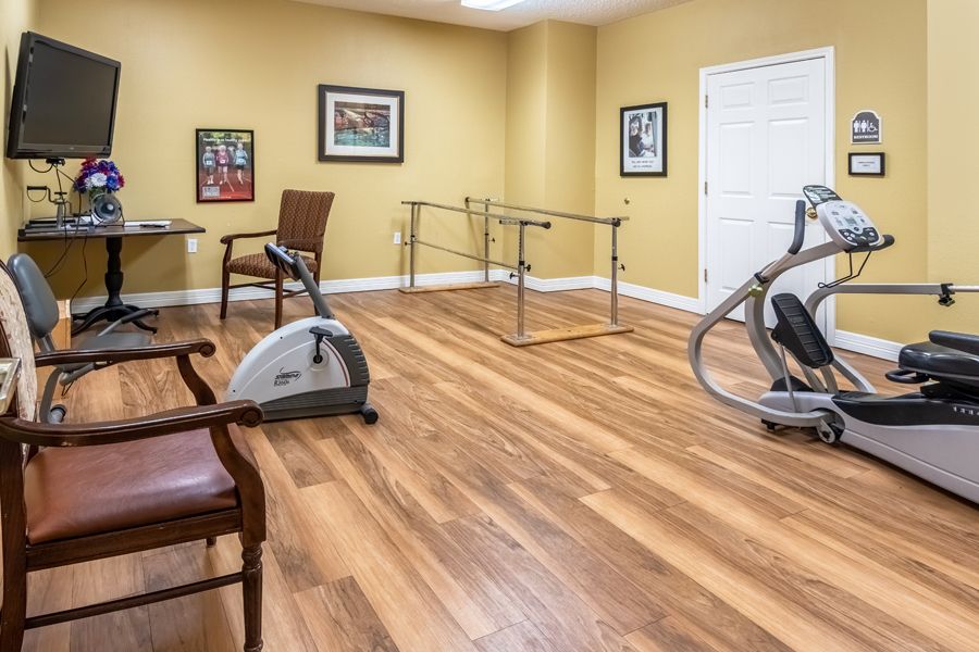 A rehabilitation exercise room featuring a stationary bike, parallel bars, and wooden flooring.