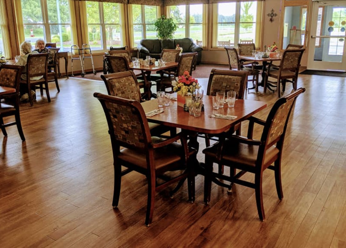 A cozy dining area with neatly arranged tables and chairs, surrounded by large windows and wooden flooring.