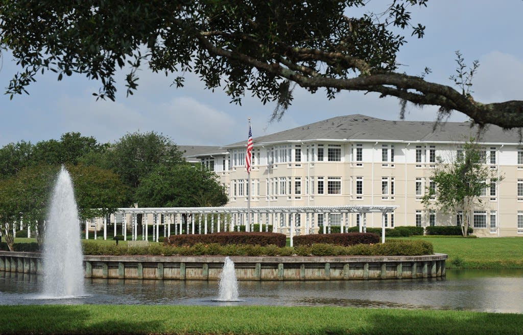 A scenic view of a large building with a pond and fountain in the foreground, surrounded by trees and featuring a flag on display.