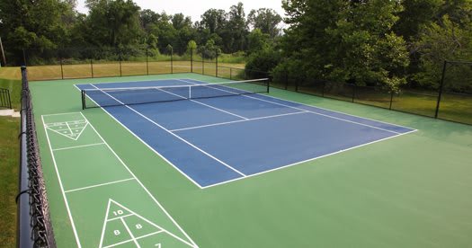 An outdoor tennis court with a blue playing surface and green borders, surrounded by nature.