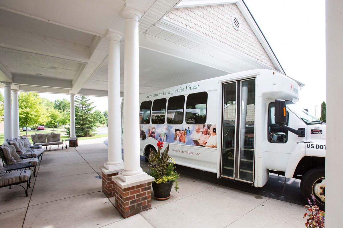 A white shuttle bus parked under a covered entrance with columns, featuring decorative plants in the foreground.